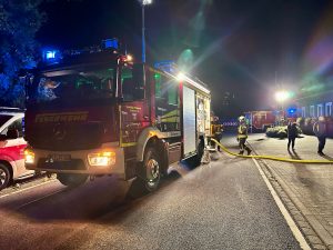 Feuerwehrfahrzeuge und Einsatzkräfte der Feuerwehr Kranenburg bei einem nächtlichen Einsatz vor einem Wohngebäude. Ein Löschfahrzeug steht mit eingeschaltetem Blaulicht auf der Straße, mehrere Schlauchleitungen sind verlegt und weitere Einsatzkräfte befinden sich im Hintergrund.